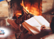 Person reading a book while sitting by a brick fireplace with a warm fire burning.